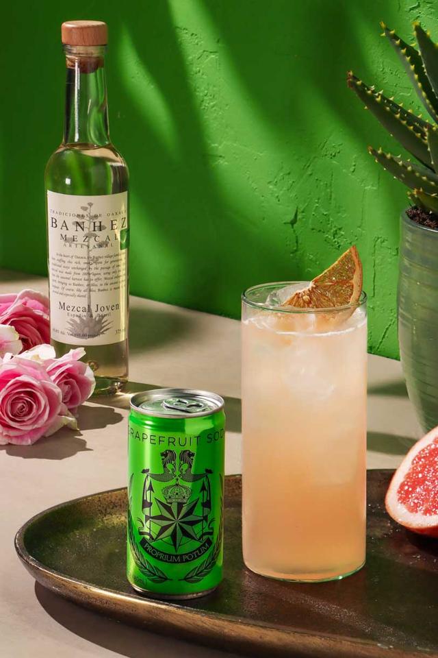 The Oaxacan Rose Paloma cocktail and a can of East Imperial grapefruit soda on the foreground with a bottle of Banhez Mezcal in the background and accents or roses, grapefruit, and an aloe plant.