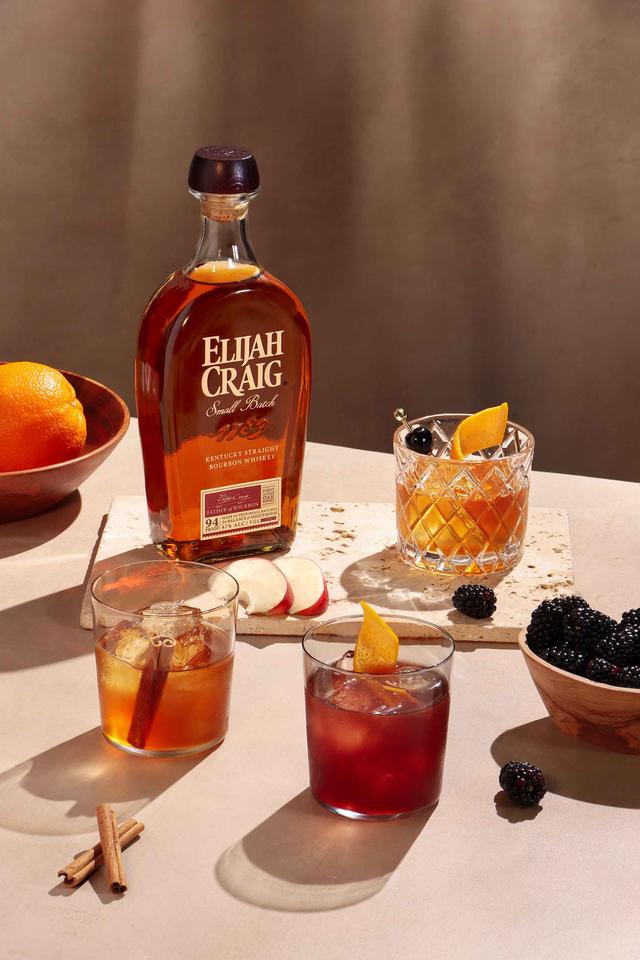 A bottle of Elijah Craig bourbon with 3 old fashioned cocktails surrounded by garnishes (orange, apple slices, cinnamon sticks, blackberries)