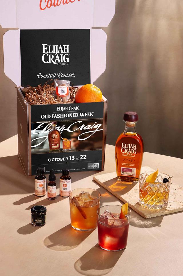 A bottle of Elijah Craig bourbon with 3 old fashioned cocktails surrounded by garnishes (orange, apple slices, cinnamon sticks, blackberries)